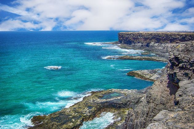 Una costa frastagliata e rocciosa costeggia un oceano turchese con piccole onde bianche sotto un cielo azzurro con nuvole soffici.