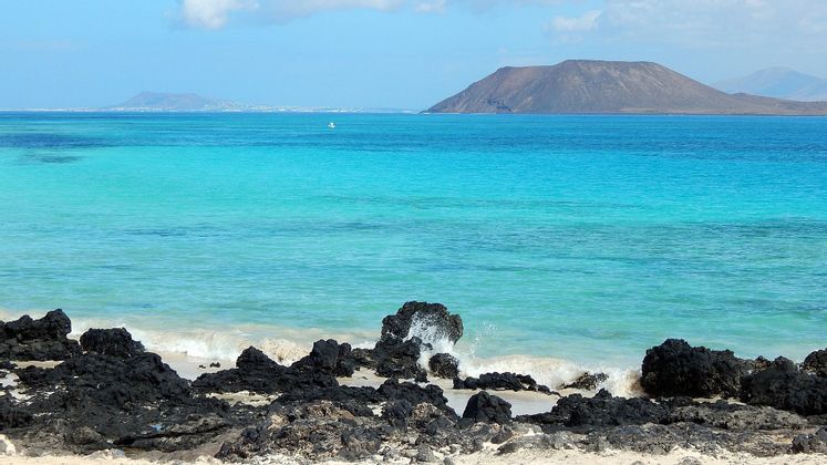 Acqua turchese lambisce rocce vulcaniche nere su una spiaggia sabbiosa, con un'isola vulcanica in lontananza sotto un cielo blu.