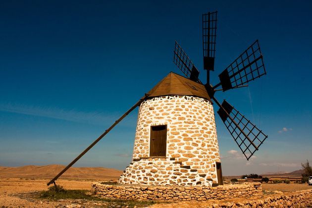 Un mulino a vento tradizionale in pietra bianca con pale di legno si erge in un paesaggio secco e arido sotto un cielo azzurro chiaro.