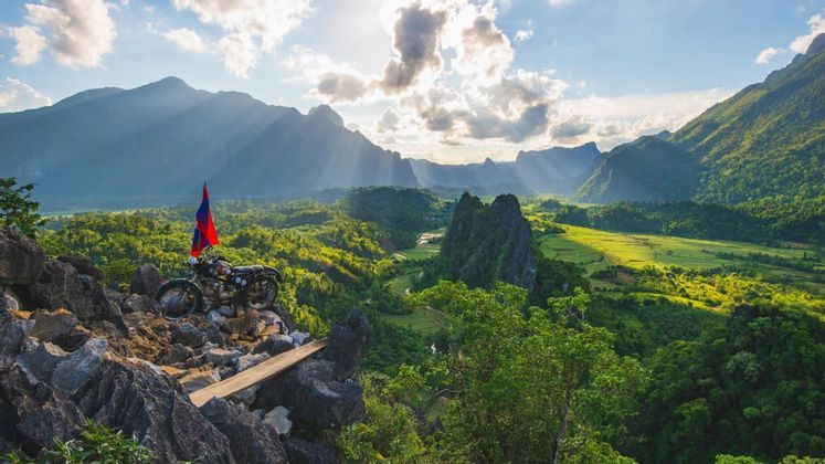 Una moto con una bandiera è parcheggiata su un belvedere roccioso che domina una lussureggiante valle montana verde, con raggi di sole che filtrano dalle nuvole.