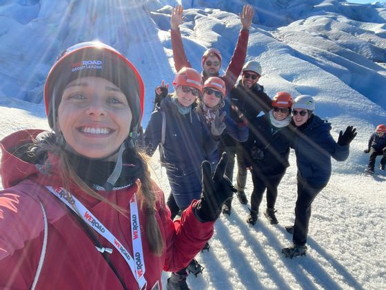 Eine WeRoad-Gruppenreiseleiterin macht ein Selfie mit ihrer Gruppe auf einem sonnenbeschienenen Gletscher, alle tragen Helme und lächeln in die Kamera.