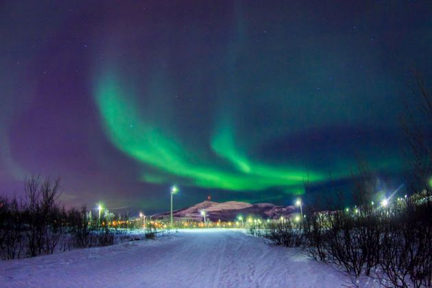 L'Aurora Boreale verde brilla in un cielo notturno stellato sopra un paesaggio innevato con lampioni e una montagna distante.