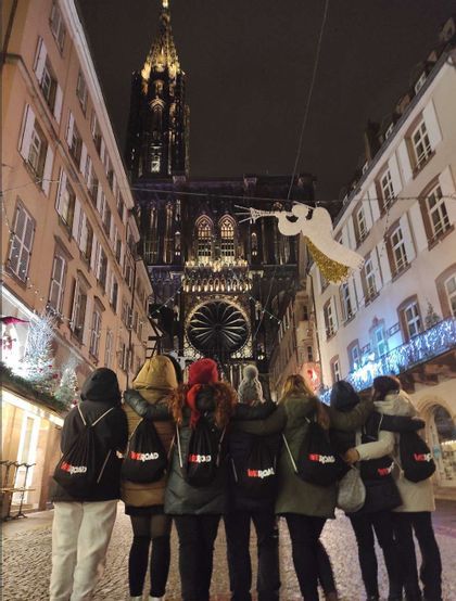 Un gruppo WeRoad in viaggio, visto di spalle, in piedi a braccetto su una strada acciottolata decorata di notte, mentre osserva una grande cattedrale illuminata.