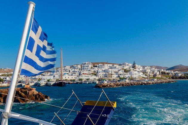 Una bandiera greca sventola su una barca che si allontana da una cittadina costiera con edifici bianchi e un porto sotto un cielo azzurro.