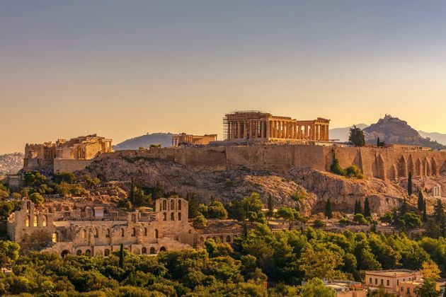 Un antico complesso di templi in pietra si erge su una collina rocciosa al tramonto, con rovine e alberi verdi sottostanti.