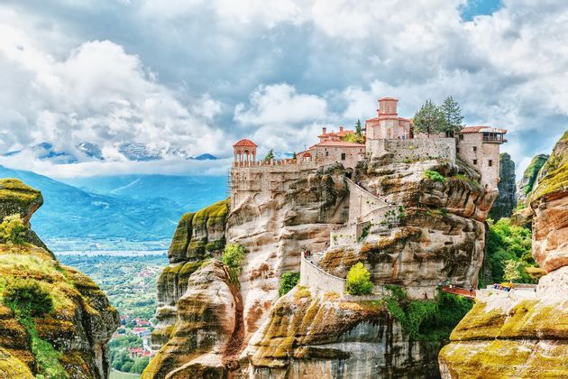 Un monastero di pietra eretto su uno sperone di roccia a picco, con una scalinata tortuosa e una vista su una vasta valle di montagna sotto un cielo nuvoloso.