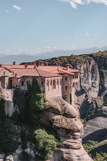 Un monastero di pietra con tetto in tegole rosse costruito sul bordo di una ripida scogliera rocciosa, con montagne distanti sullo sfondo.
