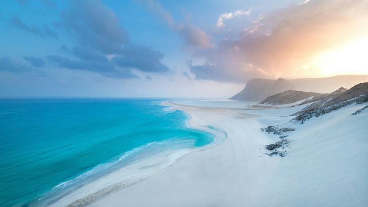 A coastline of vast white sand dunes sloping down to meet the turquoise water of the ocean under a pastel sky.