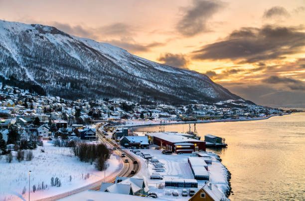 Un pueblo costero cubierto de nieve se asienta al pie de una gran montaña, con un camino serpenteante y una bahía tranquila al atardecer.
