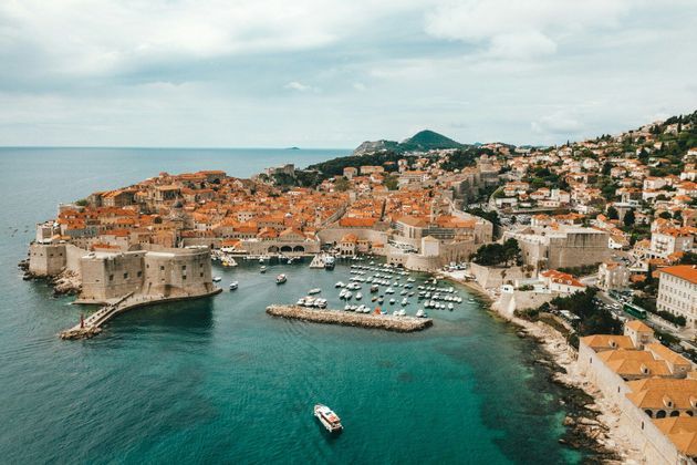 Vue aérienne d'une ville historique fortifiée aux toits de tuiles oranges sur une péninsule, avec des bateaux dans un petit port.