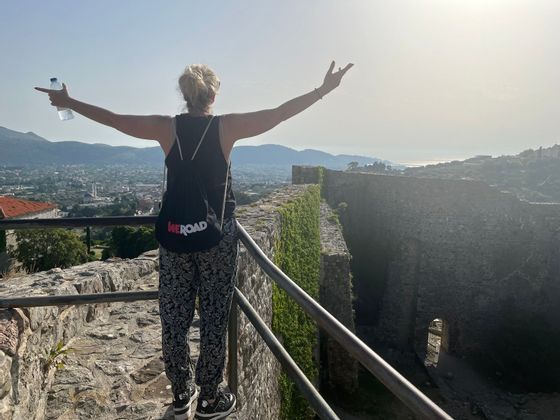 Une femme avec un sac à dos WeRoad se tient sur un mur de pierre, les bras tendus, surplombant une ville côtière depuis un point de vue élevé.