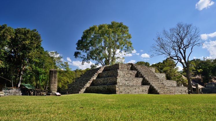 Un'antica piramide di pietra e una stele scolpita poggiano su un prato erboso, circondate da una fitta foresta sotto un cielo azzurro brillante.
