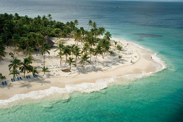 Una vista aérea de una playa tropical con arena blanca, palmeras y tumbonas, bañada por olas turquesas.