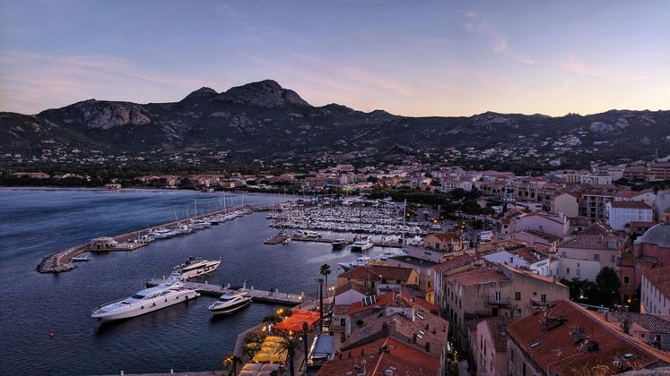 Una vista sopraelevata di un porto pieno di barche e yacht, con una città costiera e montagne sullo sfondo al tramonto.