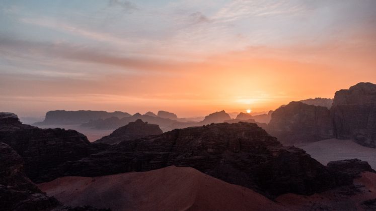 Il sole sorge sulle formazioni montuose rocciose del deserto e sulle dune di sabbia sotto un cielo arancione fosco