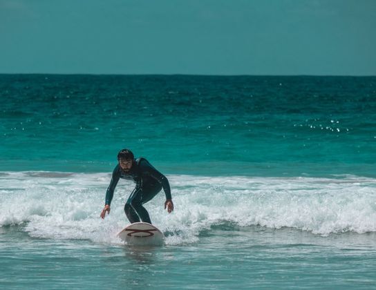 Un uomo in muta nera fa surf su una tavola bianca, cavalcando una piccola onda nel mare turchese.