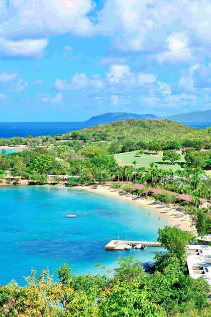Una vista dall'alto di una baia tropicale con acqua turchese, una spiaggia sabbiosa fiancheggiata da palme e lussureggianti colline verdi.