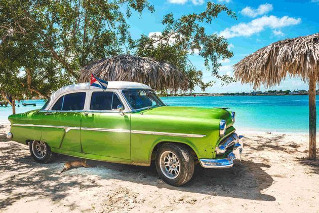 Un'auto d'epoca verde con bandiera cubana parcheggiata su una spiaggia sabbiosa accanto a un mare turchese sotto un ombrellone di paglia.