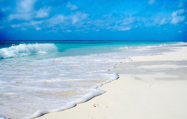 Un'onda turchese con schiuma bianca si infrange su una spiaggia di sabbia bianca sotto un cielo azzurro con nuvole soffici.