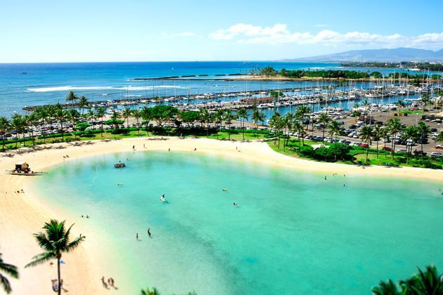 Una veduta aerea di una baia tropicale con persone che nuotano nelle acque turchesi, accanto a una spiaggia sabbiosa e un porto turistico pieno di barche.