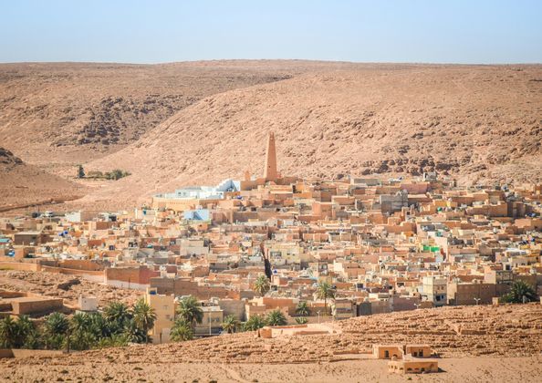Un villaggio desertico con edifici color sabbia e un minareto centrale è annidato ai piedi di una grande collina rocciosa sotto un cielo limpido.