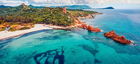 Vista aérea de una costa con agua turquesa, una playa de arena blanca, rocas rojas y colinas verdes bajo un cielo nublado.