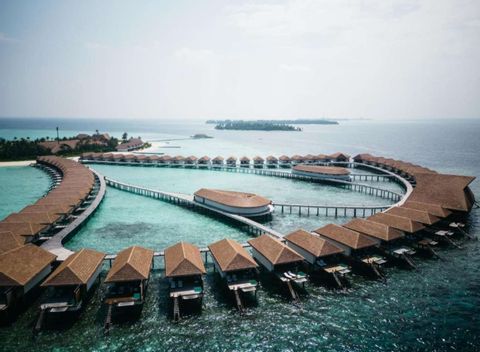 Vista aérea de un resort con bungalows sobre el agua dispuestos en curva sobre el océano turquesa.