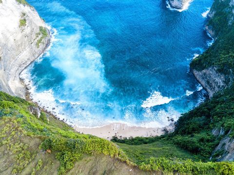 An aerial view of a secluded sandy beach in a cove, with turquoise waves crashing between steep, green cliffs.