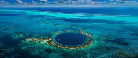 Luftaufnahme eines großen blauen Lochs, umgeben von einem Korallenriff in türkisfarbenem Wasser, mit zwei kleinen Booten, die darin fahren.