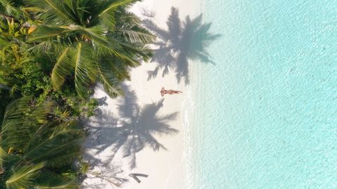 Vista aérea de una mujer relajándose en una playa de arena blanca, con sombras de palmeras y agua turquesa cristalina.