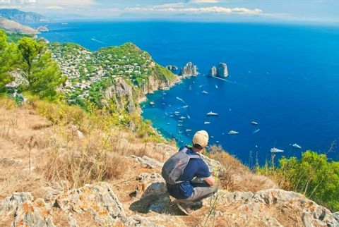 Eine Person mit Rucksack hockt auf einer felsigen Klippe und blickt auf eine Küstenstadt und ein blaues, von Booten übersätes Meer.