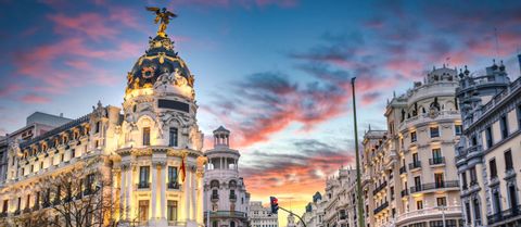 Edifici cittadini decorati, uno con tetto a cupola e statua dorata, sono illuminati contro un cielo vibrante al tramonto.