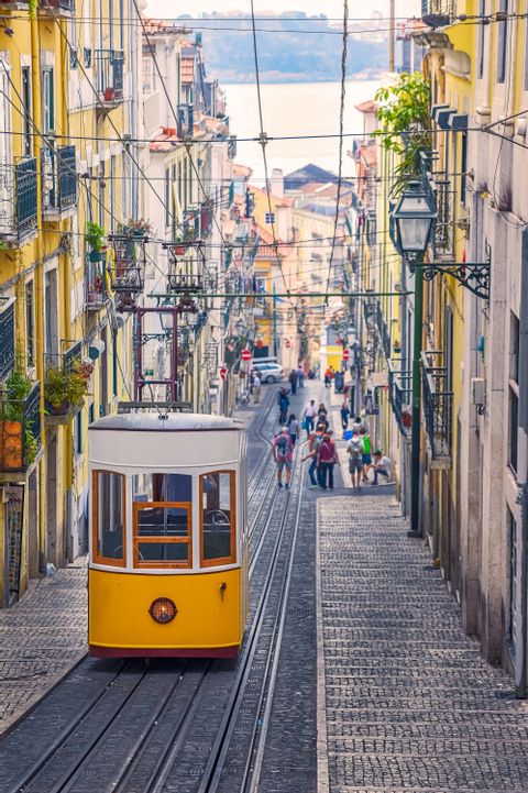 A yellow funicular tram travels up a steep, narrow cobblestone street lined with colorful buildings, with a view of the water in the distance.