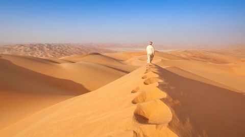 Une personne en robe claire marche le long de la crête d'une dune de sable, laissant des empreintes de pas derrière elle dans un vaste paysage désertique.