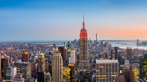 Una vista aérea del horizonte de la ciudad de Nueva York al anochecer, con el Empire State Building iluminado en rojo contra un cielo degradado.