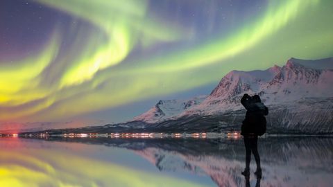 A silhouetted person photographs the green aurora borealis over snow-covered mountains, reflected in calm water at night.