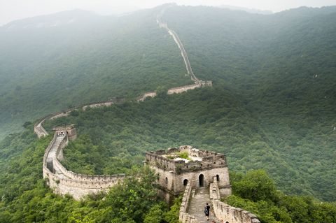 Eine Steinmauer mit Wachtürmen schlängelt sich über weite, üppig grüne Berge unter einem nebligen, bedeckten Himmel, während Menschen darauf entlangwandern.