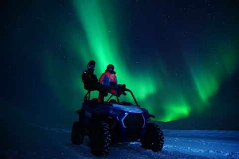 Dos personas con cascos sentadas en un vehículo todoterreno en la nieve, observando la aurora boreal iluminar el cielo nocturno estrellado.