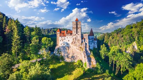 Un castillo blanco con tejados rojos se asienta sobre un acantilado rocoso rodeado por un denso bosque verde bajo un cielo azul con nubes.