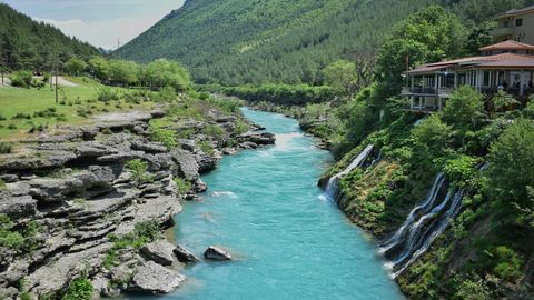 Ein türkiser Fluss schlängelt sich durch eine felsige Schlucht, wo kleine Wasserfälle einen üppigen Berghang neben einem Gebäude hinabstürzen.