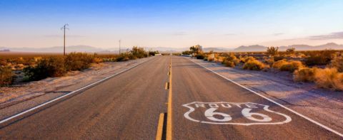 Un cartello della Route 66 dipinto sull'asfalto di un'autostrada vuota che attraversa un paesaggio desertico con montagne in lontananza.