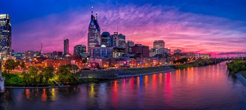 La silhouette illuminée d'une ville se reflète dans une large rivière au coucher du soleil, sous un ciel rose et violet éclatant.
