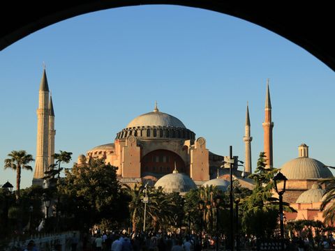 Una gran mezquita con múltiples cúpulas y minaretes, enmarcada por un oscuro arco contra un cielo azul claro al atardecer.