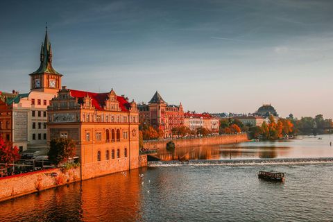 Historic buildings with red roofs and a clock tower line a river during a golden sunset, with a tour boat in the water.