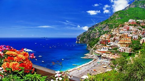 Un pueblo colorido en un acantilado verde con vistas a un mar azul salpicado de barcos, con flores brillantes en primer plano.