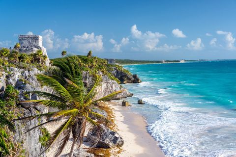 Ancient stone ruins sit atop a rocky, jungle-covered cliff overlooking a white sand beach and turquoise ocean.