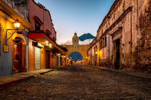 Una calle empedrada vacía al anochecer conduce a un arco amarillo con una torre del reloj, con un volcán visible a lo lejos.