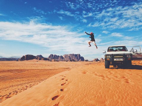 A person jumps in the air on an orange sand dune in the desert, next to a white pickup truck, with mountains in the background.
