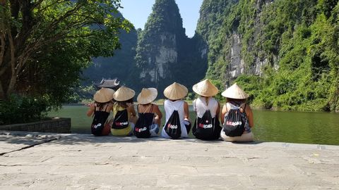 Un groupe WeRoad est assis sur une corniche rocheuse, coiffé de chapeaux coniques et admirant une rivière entourée de montagnes verdoyantes.