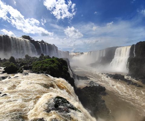 Inmensas cascadas caen sobre acantilados rocosos y exuberantes hacia un cañón neblinoso bajo un cielo azul con nubes blancas.